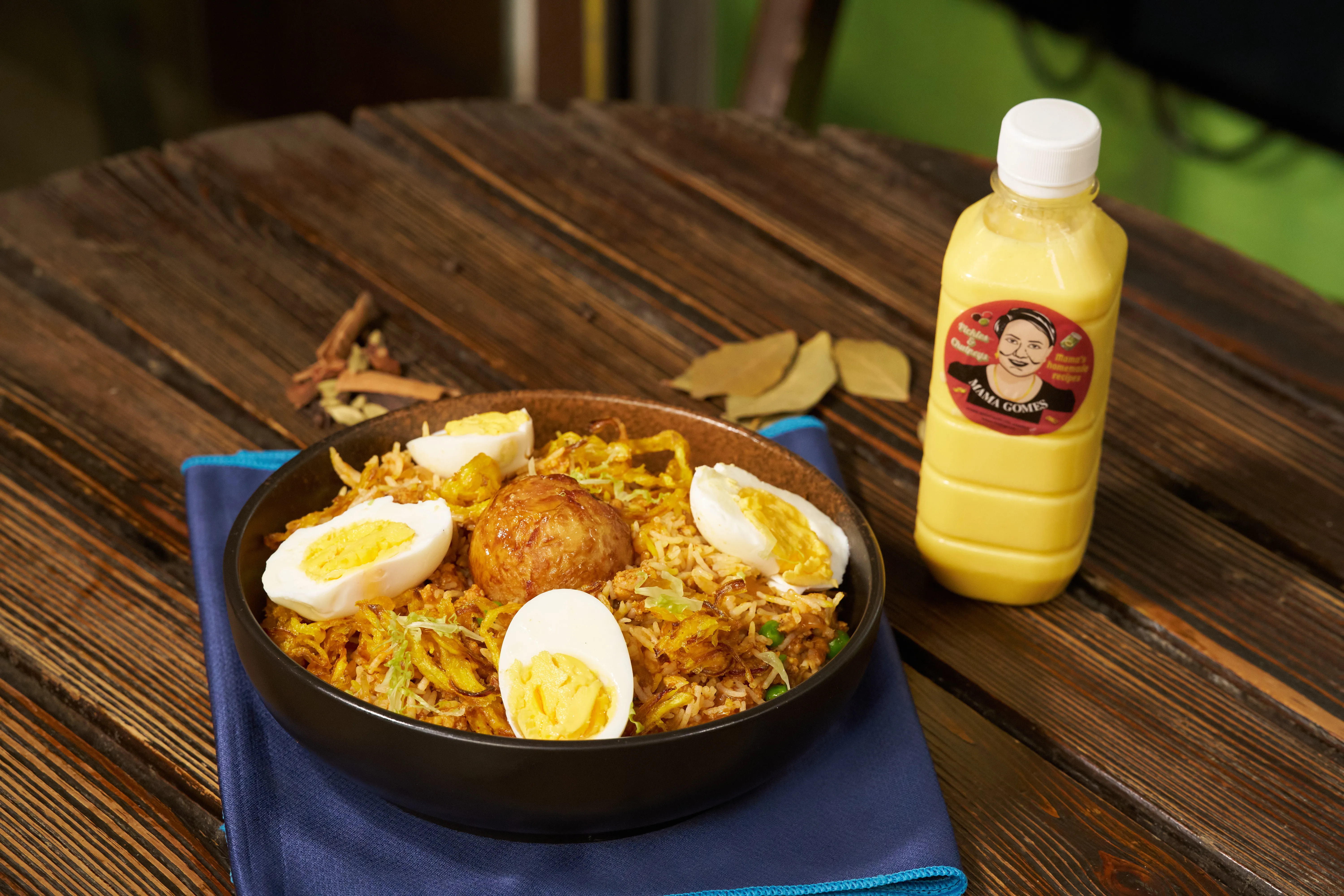 A bowl of biryani with boiled eggs and a bottle of Mama Gomes lassi on the wooden street table