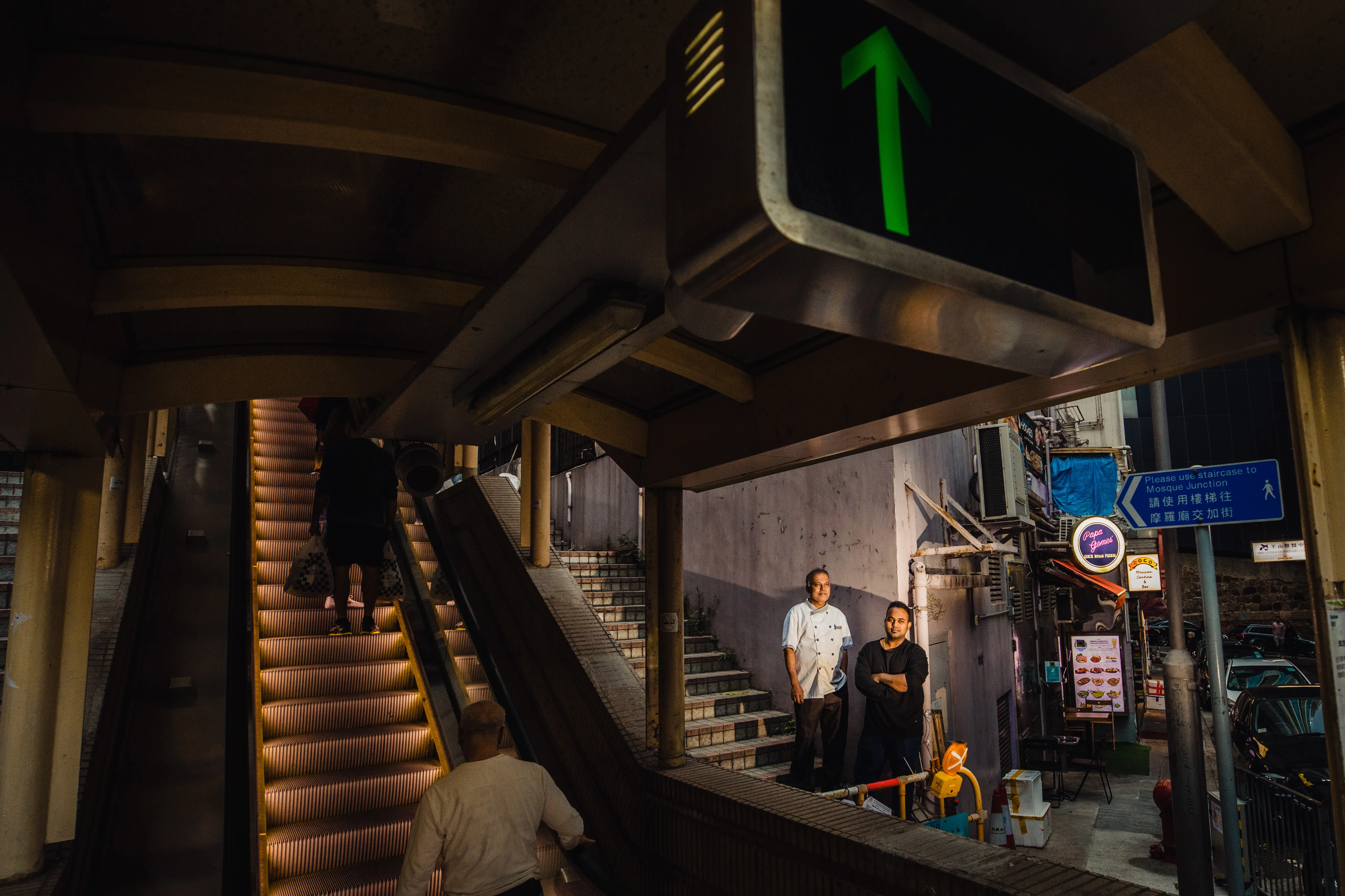 Monu and Alex Gomes standing at the foot of the Central–Mid-Levels Escalator beside the lit Papa Gomes sign