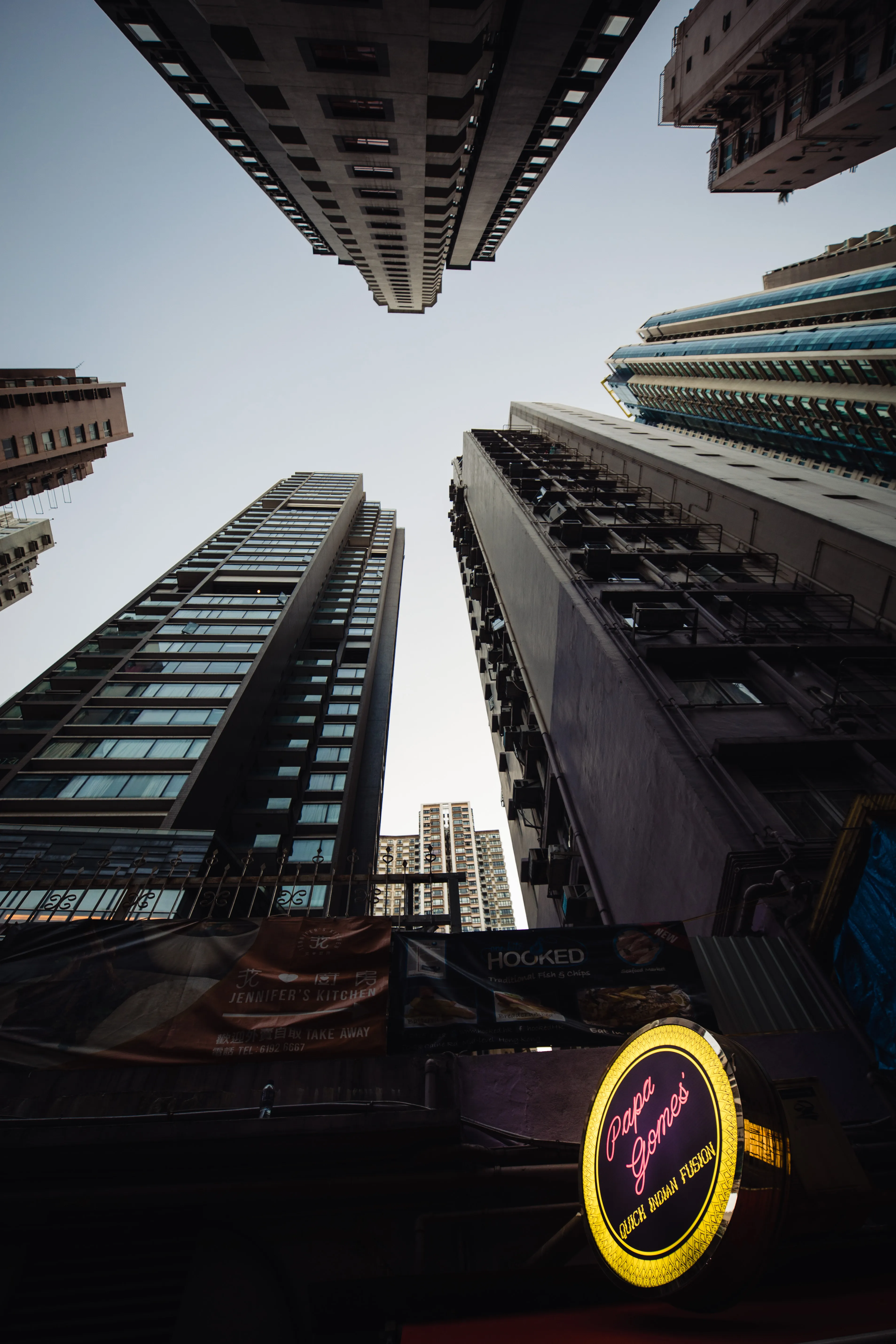 The illuminated Papa Gomes badge sign on Mosque Street at dusk, seen from below against the residential towers of Mid-Levels