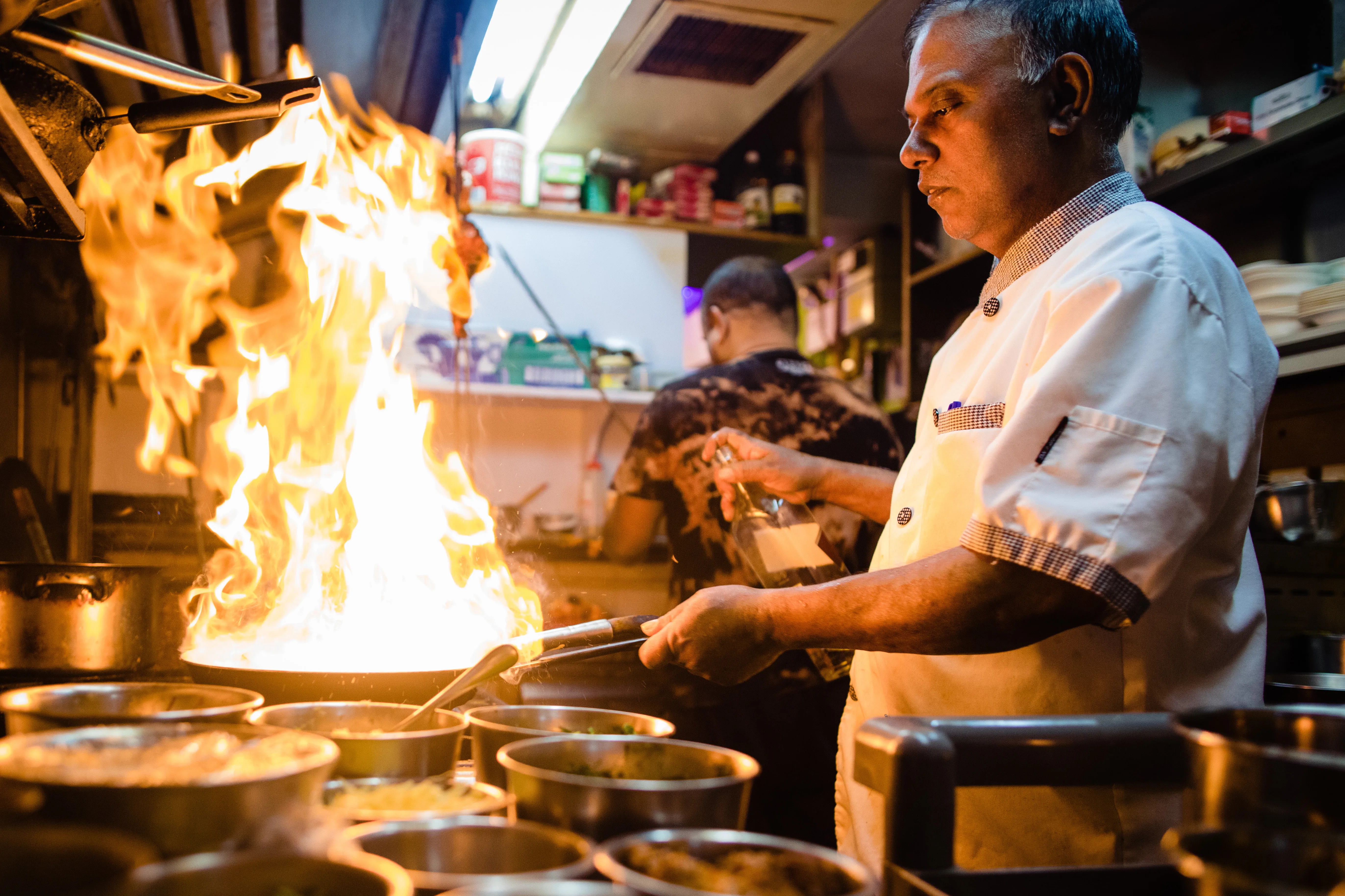 Monu Gomes at the pass, flames rising from the pan, tandoori skewers hanging behind him
